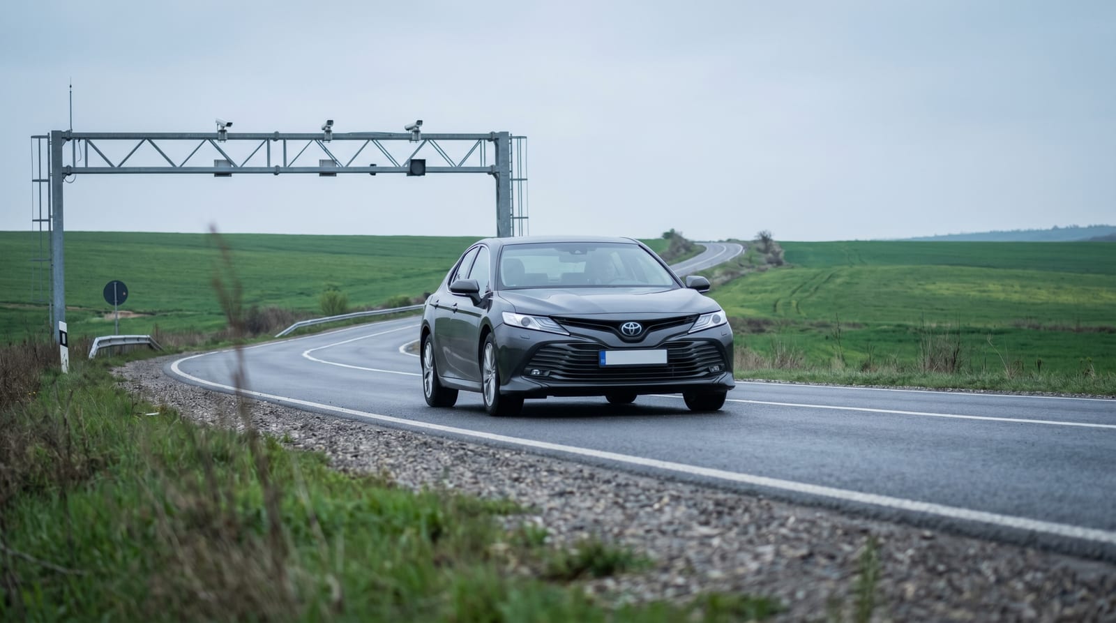 Autoturism trecând pe sub un portal cu camere de monitorizare a rovinietei, pe un drum național printre câmpuri, într-o zi cu cer acoperit