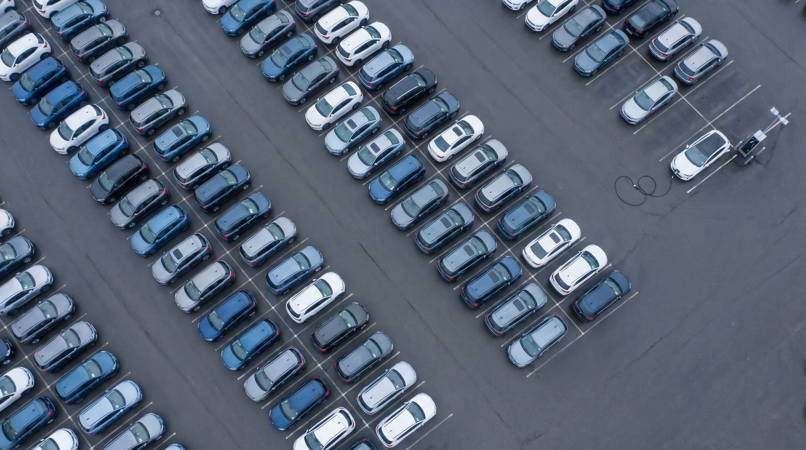Parcarea unui dealer auto văzută de sus, cu zeci de mașini noi aliniate pe rânduri și o singură stație de încărcare cu cablul conectat